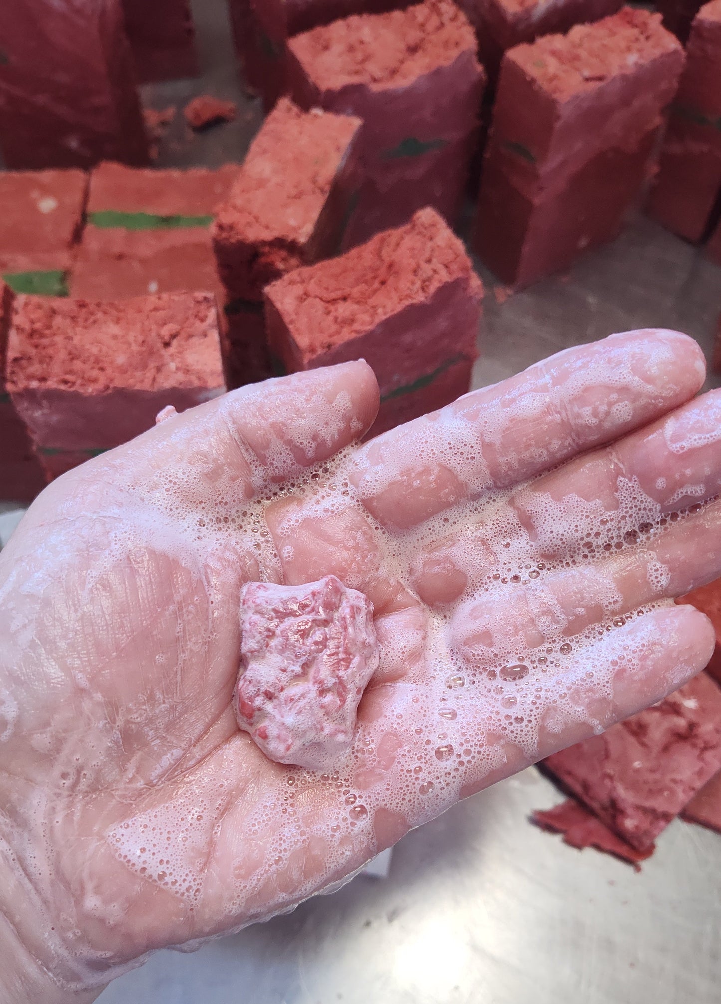 Hand with soap suds holding a small block of red soap next to stacked red soap bars.